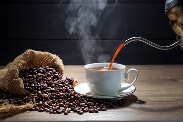 Pouring hot coffee into a white coffee cup With roasted coffee beans on a wooden table, dark black background.