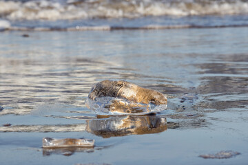 
chunks of ice on a gray sandy surface with reflections, water and waves in the background.