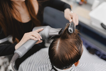 Female hairstylist drying and styling hair of young caucasian male at barbershop, warm toning