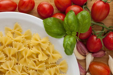plate of durum wheat pasta, farfalle shape, on wooden cutting board with ripe tomatoes, fresh basil leaves and garlic cloves