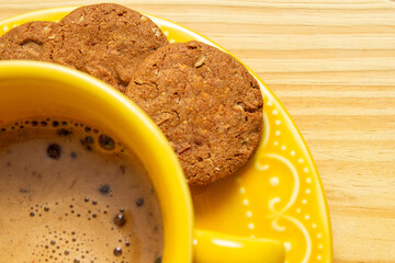 Detalhe de uma xícara amarela com cappuccino e alguns cookies no píres sobre superfície de madeira.