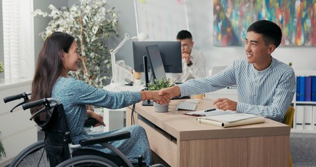 Woman is sitting in wheelchair at desk in office across employer they are signing employee contract...