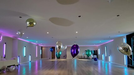 Start with the wide living room illuminated by deep purple and turquoise neon lights. Balloons in shades of black and silver descend slowly. - Powered by Adobe