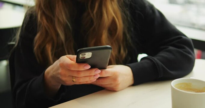 WARSAW, POLAND - MARCH 10 2023: Woman with wavy blonde hair holds phone and looks at screen sitting at table. Warm cup of coffee stays nearby as lady enjoys free time in cozy cafe