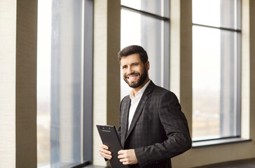 Young smiling attractive entrepreneur in formal suit standing in modern office. Real estate agent man holding documents and looking cheerful at camera. Business portrait and real estate concept.