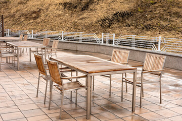 Empty outdoor café setting with wooden tables and chairs in early spring. The tables are rectangular and have a slatted top, while the chairs have a similar slatted design.