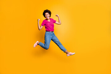 Full length photo of crazy ecstatic girl raise her fists scream shout yeah celebrate victory lottery discounts jump wear red t-shirt trendy denim jeans sneakers isolated over yellow color background