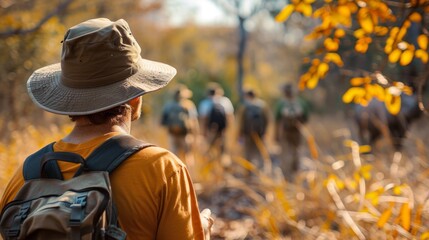A person wearing a hat and backpack observes a group of hikers on a trail, surrounded by tall grass and colorful autumn leaves in the afternoon sunlight.