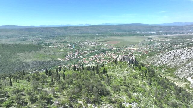 AERIAL: Stjepan Grad fortress standing on a rocky hill above town of Blagaj. Medieval fortress amidst lush greenery and mountain ridges with views of the inhabited valley and winding Buna River below.