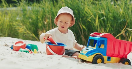 Boy on the beach near the water pours sand into a bucket with a shovel, surrounded by toys, deeply...