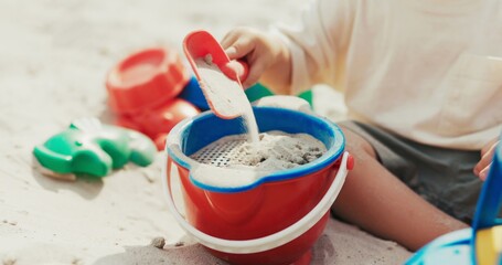 Boy plays in the sandbox, pouring sand from a shovel into a bucket, building castles on his own in the backyard.