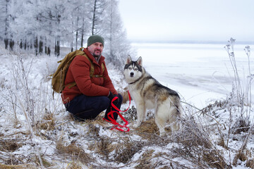 A beautiful blue-eyed husky and his happy male owner, dressed in a red jacket and backpack, during a winter walk along the shore of a frozen lake