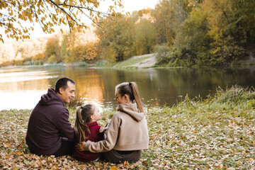 Naklejka premium Happy family sitting under tree over lake in park outdoors. Autumn season.