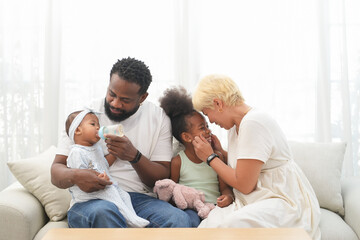 Obraz premium Loving interracial family sitting on couch at home,an african father take care a baby,feeding milk and asian mother playing with a girl