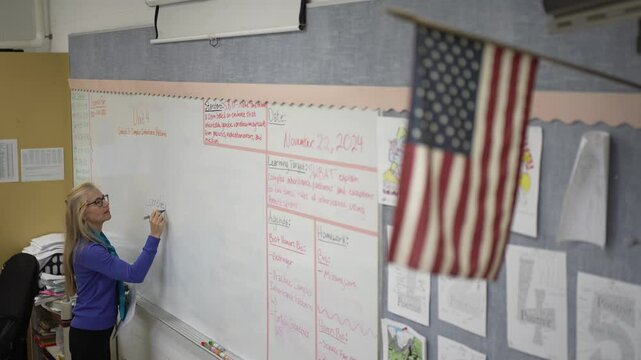 A middle-aged woman stands in front of a whiteboard, writing important notes for her class while students look on attentively. The classroom is bright and organized.