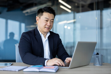 Smiling young successful Asian male businessman and office worker sitting and working at desk using laptop