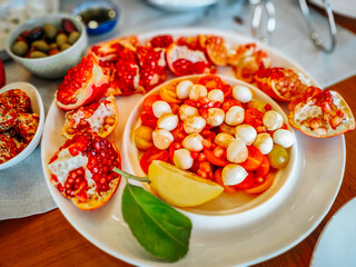 A vibrant salad with mozzarella, cherry tomatoes, grapes, and pomegranate seeds, garnished with lemon and served on a white plate, creating a colorful and refreshing appetizer.