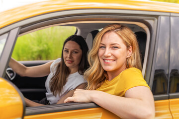 A smiling blonde woman leans out of a yellow taxi's window while her brunette friend sits in the driver's seat, both enjoying their journey.