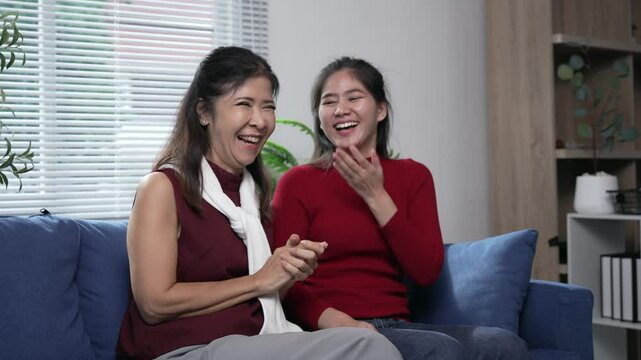 Mother and daughter are sitting on a blue sofa in the living room, laughing and having fun together, sharing a moment of joy and complicity