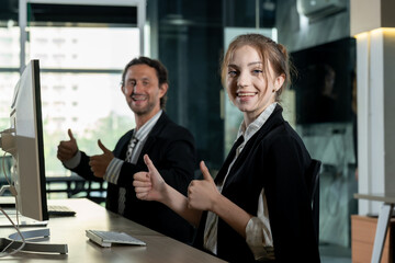 Smiling businesswoman and businessman sitting at office desk showing thumbs up. Both wearing formal business attire. Large office window revealing cityscape background. Positive teamwork