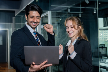 Excited businessman and businesswoman celebrating success in modern office. Holding laptop while smiling and raising fists. Glass partitions and bright lighting creating corporate achievement