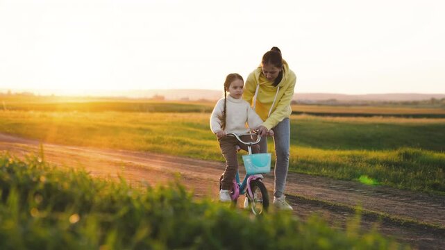 A young girl is riding a bike with her mother. The mother is holding the girl's hand and guiding her. The scene is peaceful and happy, with the sun shining down on the grassy field