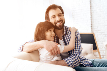 Bearded father embraces with teenager son sitting on the couch.