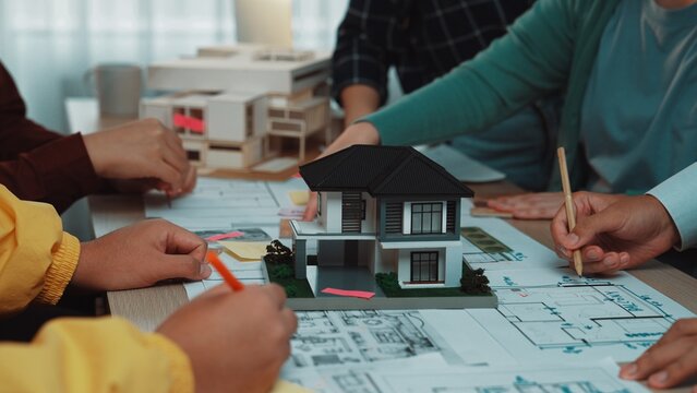 A group of architects collaborates in a modern office environment, using a scale model and floor plans to bring innovative housing ideas to life. SACTR