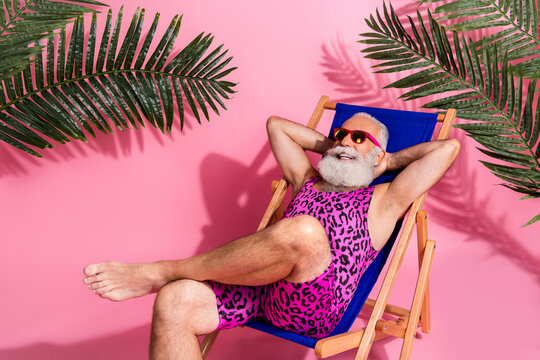 Photo of positive senior man wear swimwear hold arms behind head relax rest chaise lounge isolated on pink color background
