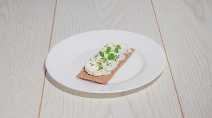 A crispbread with cream cheese, flax seeds, and microgreens on a white plate with a light wooden background.