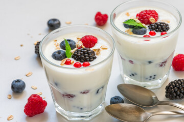 Greek yogurt with fresh berries, oats and mint in glasses on white background with spoons. Yoghurt with blueberry, raspberry, blackberry and granola. Healthy breakfast. Diet food