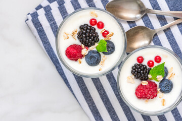 Greek yogurt with fresh berries, oats and mint in glasses on napkin over white background with spoons. Top view. Yoghurt with blueberry, raspberry, blackberry and granola. Healthy breakfast. Diet food