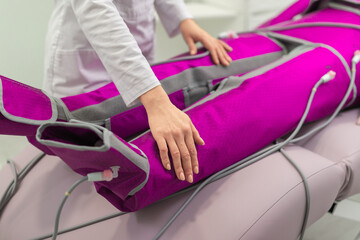 A woman undergoes a pressotherapy session, wearing a specialized purple compression suit, assisted by a therapist in a clinical setting.