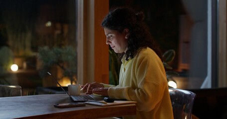 The woman works from home at night. She uses a laptop and writes in a notebook while sitting at her desk.
