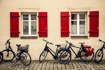 Fototapeta premium A picturesque European facade with two windows featuring red wooden shutters. Parked bicycles along the cobblestone street enhance the old-town atmosphere, ideal for travel and cityscape themes