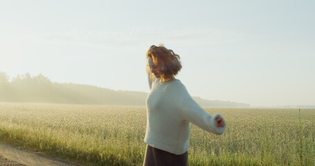 A beautiful young red-haired woman laughs as she spins and dances on a gravel road near an agricultural field in the rays of the rising sun. 4k, video, red komodo