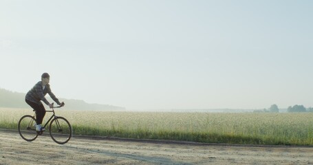 Obraz premium A young stylishly dressed man, with a camera hanging around his neck, rides a bicycle on a gravel road near a wheat field in the rays of the rising sun. 4k video, red komodo