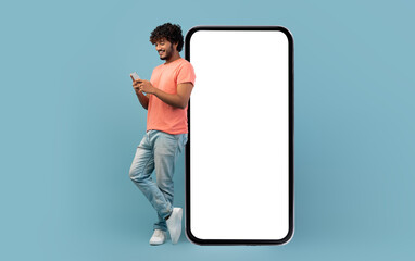 Cool curly indian guy in casual outfit leaning on huge smartphone with white blank screen, using cell phone and smiling, blue studio background, mockup, panorama with copy space