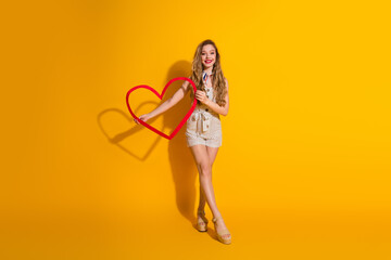Joyful young woman holding a heart-shaped frame against a bright yellow background, expressing happiness, positivity, and love