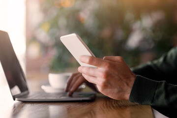 Cropped of young man holding smartphone and using laptop, cafe interior