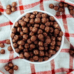 Bowl of chocolate cereal balls on a checkered cloth. Crunchy breakfast food made of cocoa-flavored puffed grains.