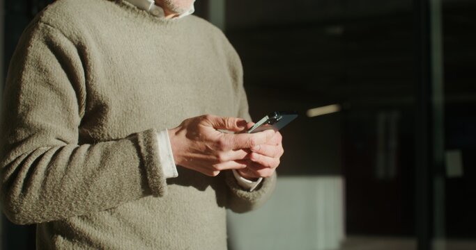 A gray-haired man dressed in sweater is typing a message on his phone during walking around the city. Close-up, the video moves from the man's hands to his face. Video in 4k, red komodo