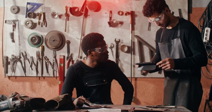 A male blacksmith in work clothes shows his colleague an idea for a sketch on a clipboard, working together to design a future product in a workshop full of tools. video in 4k, red komodo