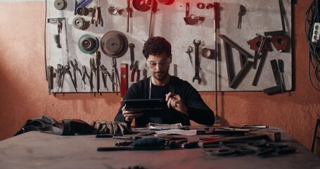 A young blacksmith in goggles and an apron uses a tablet while sketching a future product, sitting at a desk in a forge full of working tools. video in 4k, red komodo