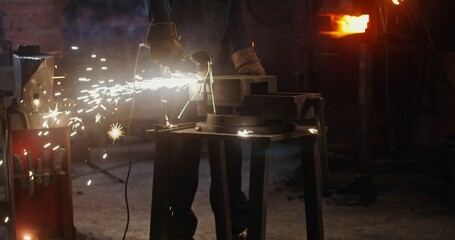 A young blacksmith of African American appearance in goggles and work clothes uses an angle grinder while working in a modern forge. Metal sparks fly in all directions. 4k, red komodo