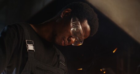 A young blacksmith of African American appearance in goggles and work clothes uses an angle grinder...