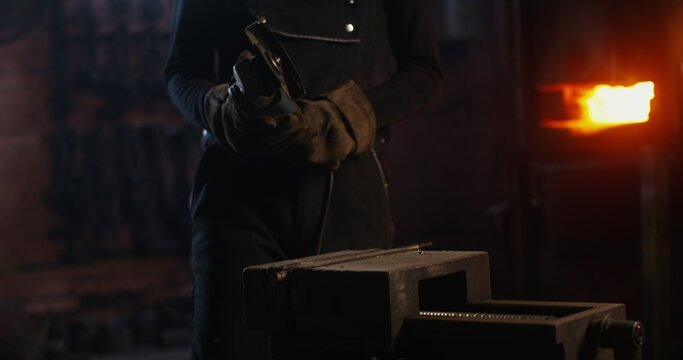 A young blacksmith of African American appearance in goggles and an apron turns on an angle grinder and begins to cut off a piece of metal workpiece while working in a forge. 4k, red komodo