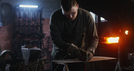 A young man of European appearance, dressed in work clothes and goggles, cuts a sheet of iron with...