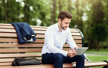 Fototapeta premium Handsome company employee relaxing on bench at park and browsing internet on tablet computer
