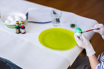 Child carefully painting Easter egg with brush while wearing gloves at decorated table with colors and raw eggs.
